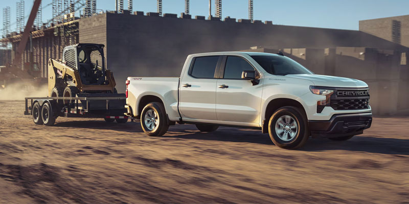 A white 2026 Chevrolet Silverado pickup truck towing a trailer with a small construction vehicle on a dusty construction site.