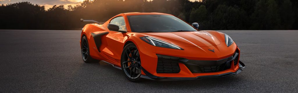 An orange 2026 Chevrolet Corvette is parked on a paved surface with trees and sunset in the background.