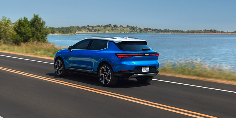 Blue 2026 Chevrolet Equinox EV driving on a scenic road beside a lake, showcasing rear design, sleek LED taillights, and modern crossover styling