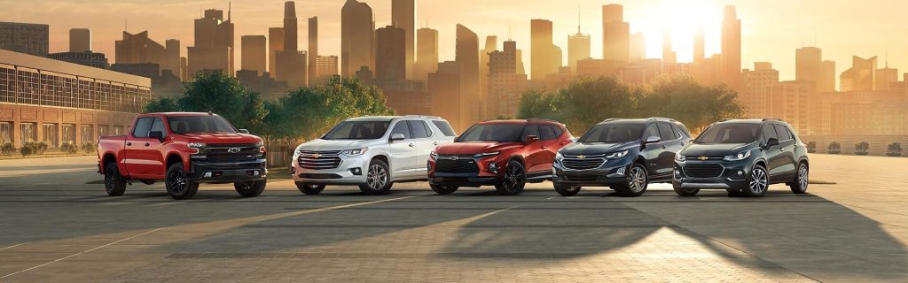 Chevrolet truck and SUV lineup parked in front of a city skyline at sunset, showcasing multiple models in a clean urban setting