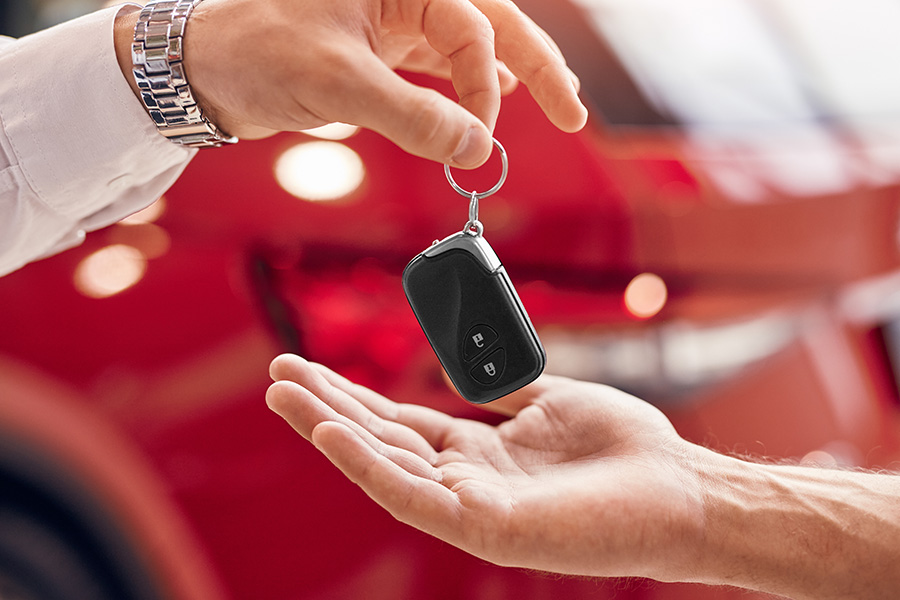 Close-up of a car key fob being handed over, symbolizing vehicle handoff or purchase completion, with a red car blurred in the background.