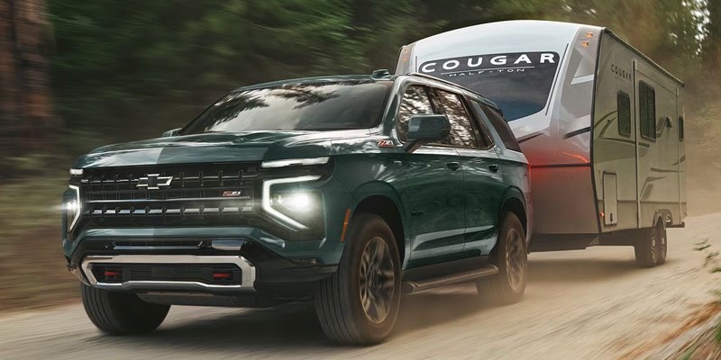 A dark green 2026 Chevrolet Tahoe towing a silver and red travel trailer on a dirt road surrounded by trees.