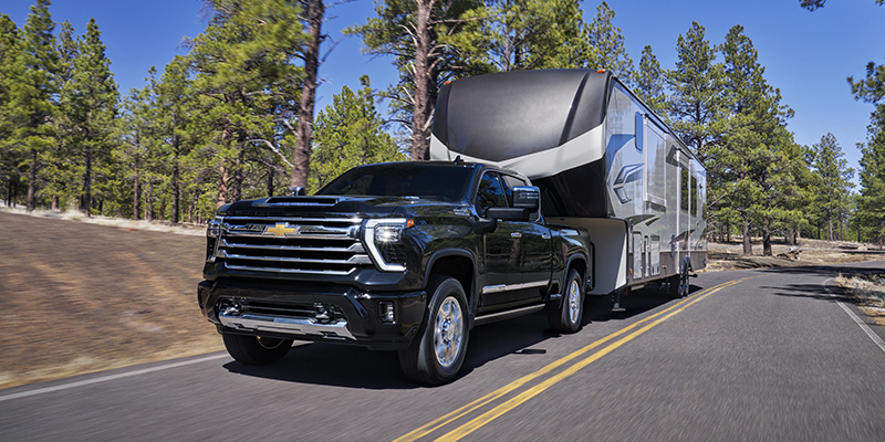 Black Chevrolet Silverado heavy-duty pickup truck towing a large fifth-wheel RV trailer along a scenic forest road on a sunny day.