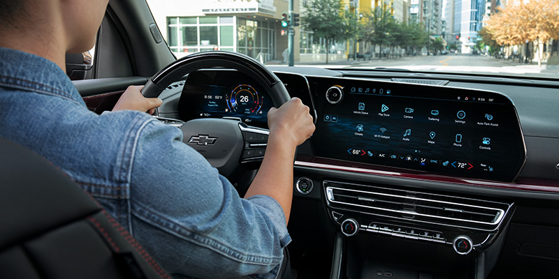 Driver behind the wheel of a Chevrolet Traverse vehicle, viewing a digital instrument cluster and large touchscreen infotainment system while driving through a city street.