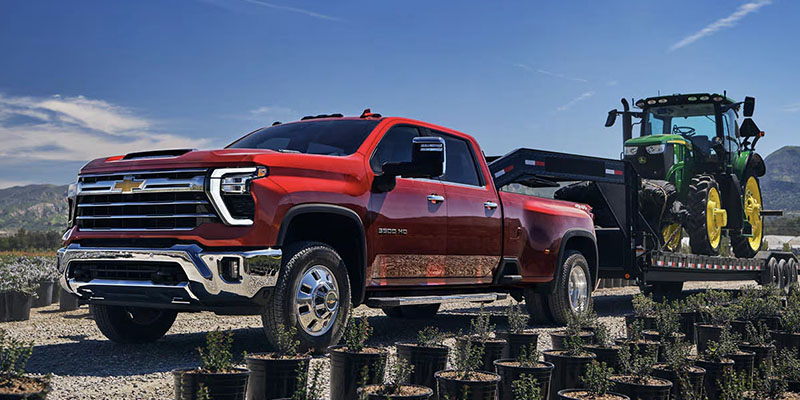 Red Chevrolet Silverado HD towing a trailer with a green tractor through a rural landscape.