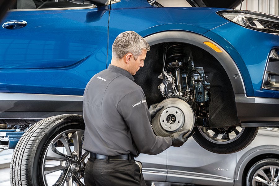 A mechanic in a gray uniform is working on the brakes of a blue Chevrolet car that is lifted up, revealing its undercarriage.