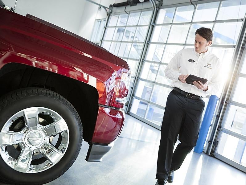 A man in a white dress shirt and black pants standing next to a red truck in a garage, holding a tablet.