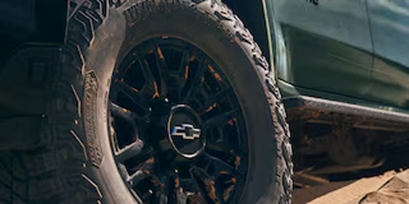 A close-up of a black wheel with a Chevrolet logo, mounted on a rugged tire, parked on dirt or gravel.
