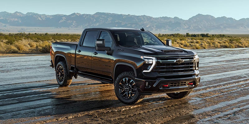 A black Chevrolet Silverado pickup truck parked on a rugged, muddy terrain with mountains in the background under a clear sky.